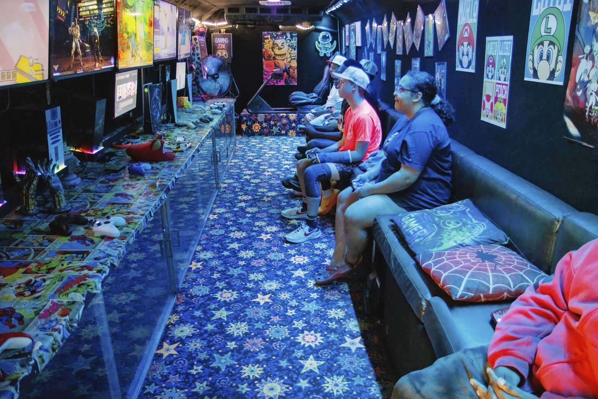 Arcade gaming room with neon lights, patterned carpet, and people seated on couches playing games