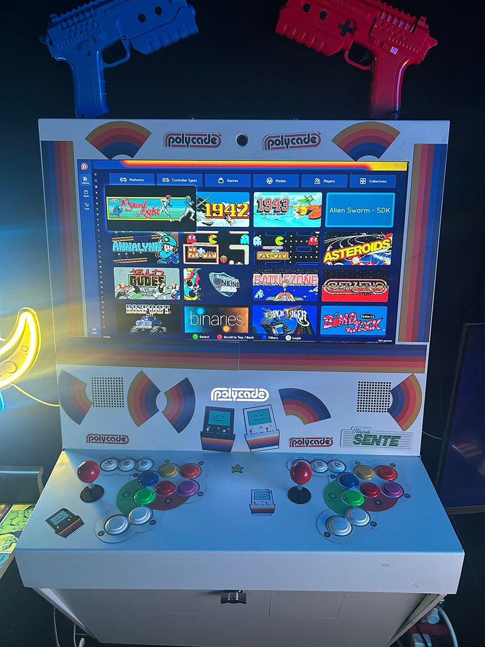 Arcade cabinet with a game selection screen, two joysticks, colorful buttons, and blue and red light guns on top.