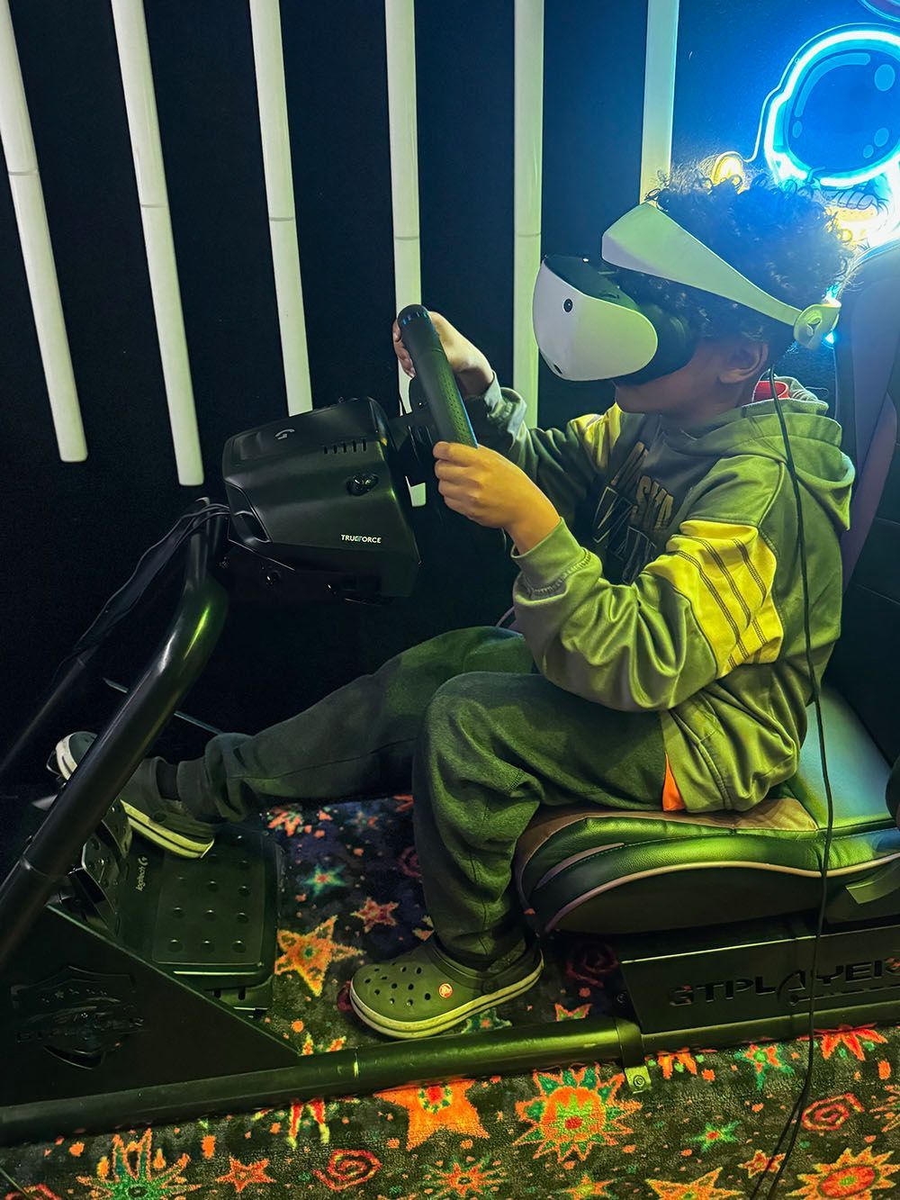 A person wearing a VR headset sits in a racing simulator chair, gripping a steering wheel in a dimly lit, patterned room.