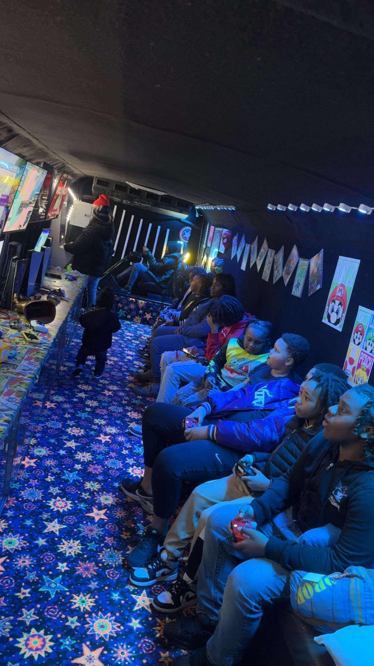 A group of people seated in a darkened, carpeted room with neon star patterns, watching a screen at the front.