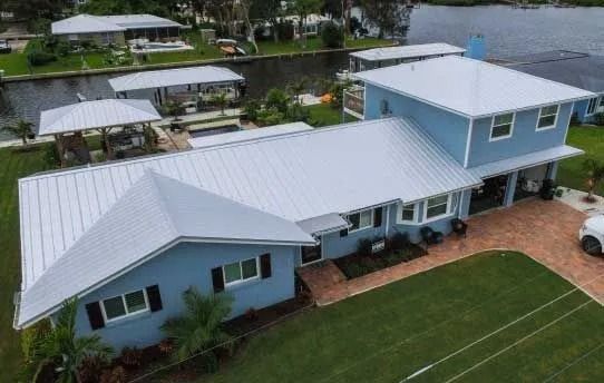 Blue house with white metal roof near water; green lawn and brick driveway.