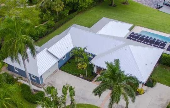 Aerial view of a modern blue house with a white metal roof, surrounded by palm trees and green grass.