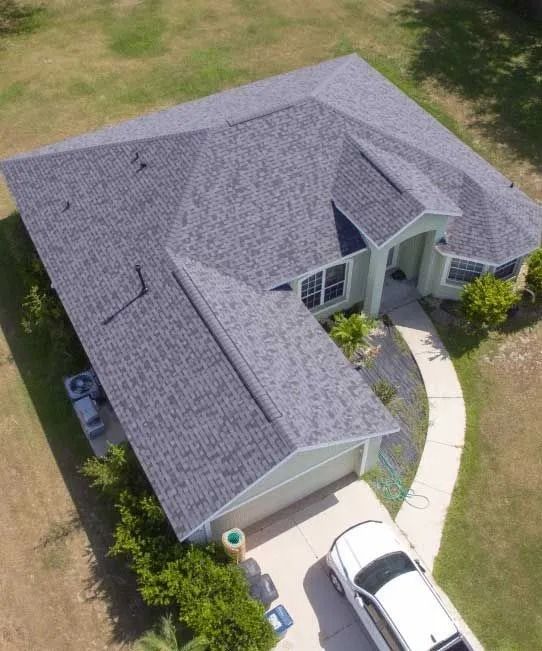 Aerial view of a light green house with a gray shingled roof, driveway, and white car.
