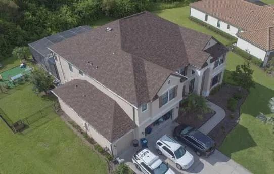 Aerial view of a two-story beige house with a brown roof, two cars in the driveway, and a screened-in pool.