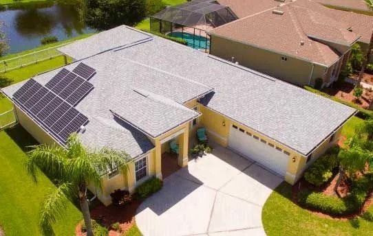 Aerial view of a yellow house with solar panels on the roof, a white garage, and a paved driveway.