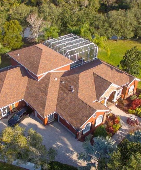Aerial view of a red-roofed house with a screened-in pool, surrounded by lush green trees and landscaping.