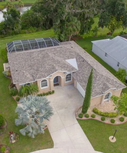 Aerial view of a beige house with a long driveway, green lawn, and a screened-in patio.