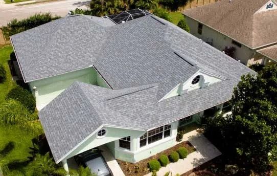 Aerial view of a light green house with a gray shingle roof and lush green landscaping.