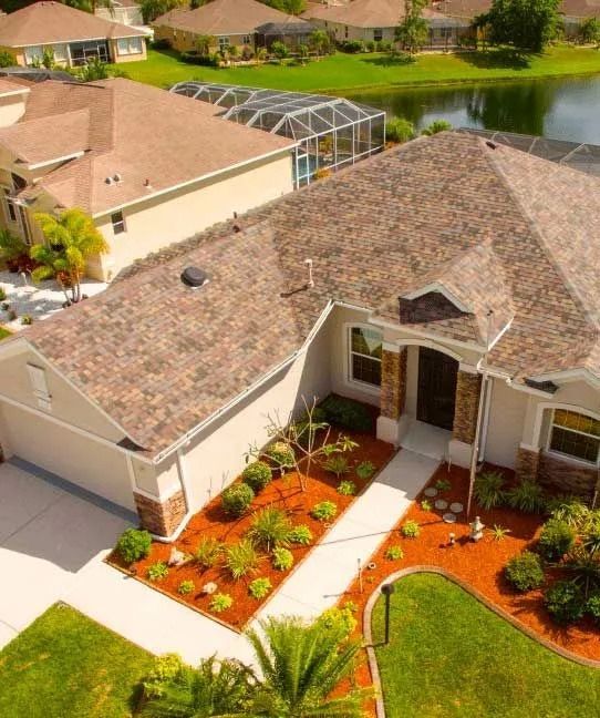 Aerial view of a house with a brown roof and beige exterior, landscaped front yard, and a lake in the background.
