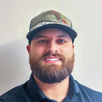 Man with beard wearing a camouflage cap and black polo shirt smiling at the camera.