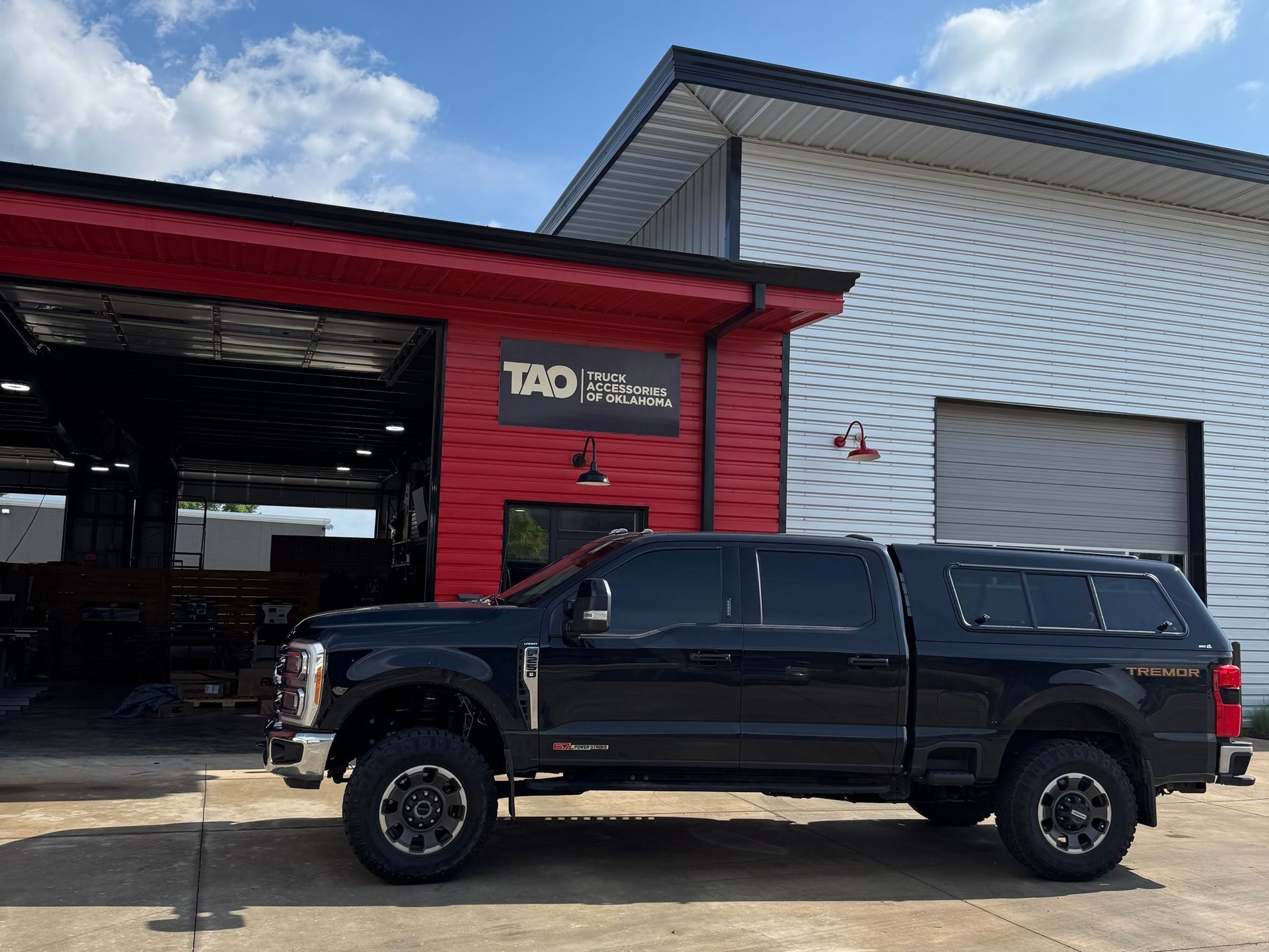 Black pickup truck with a camper shell parked outside a red and white building with a large garage door.