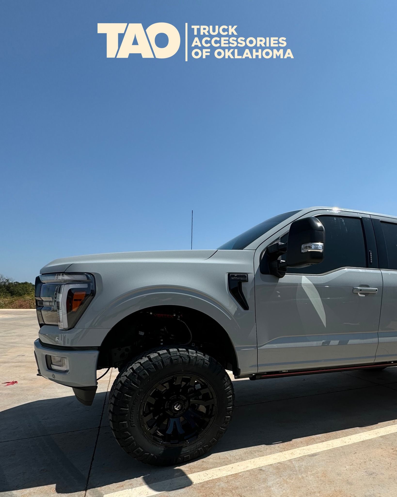 Gray lifted truck with black wheels under a blue sky; Truck Accessories of Oklahoma logo.