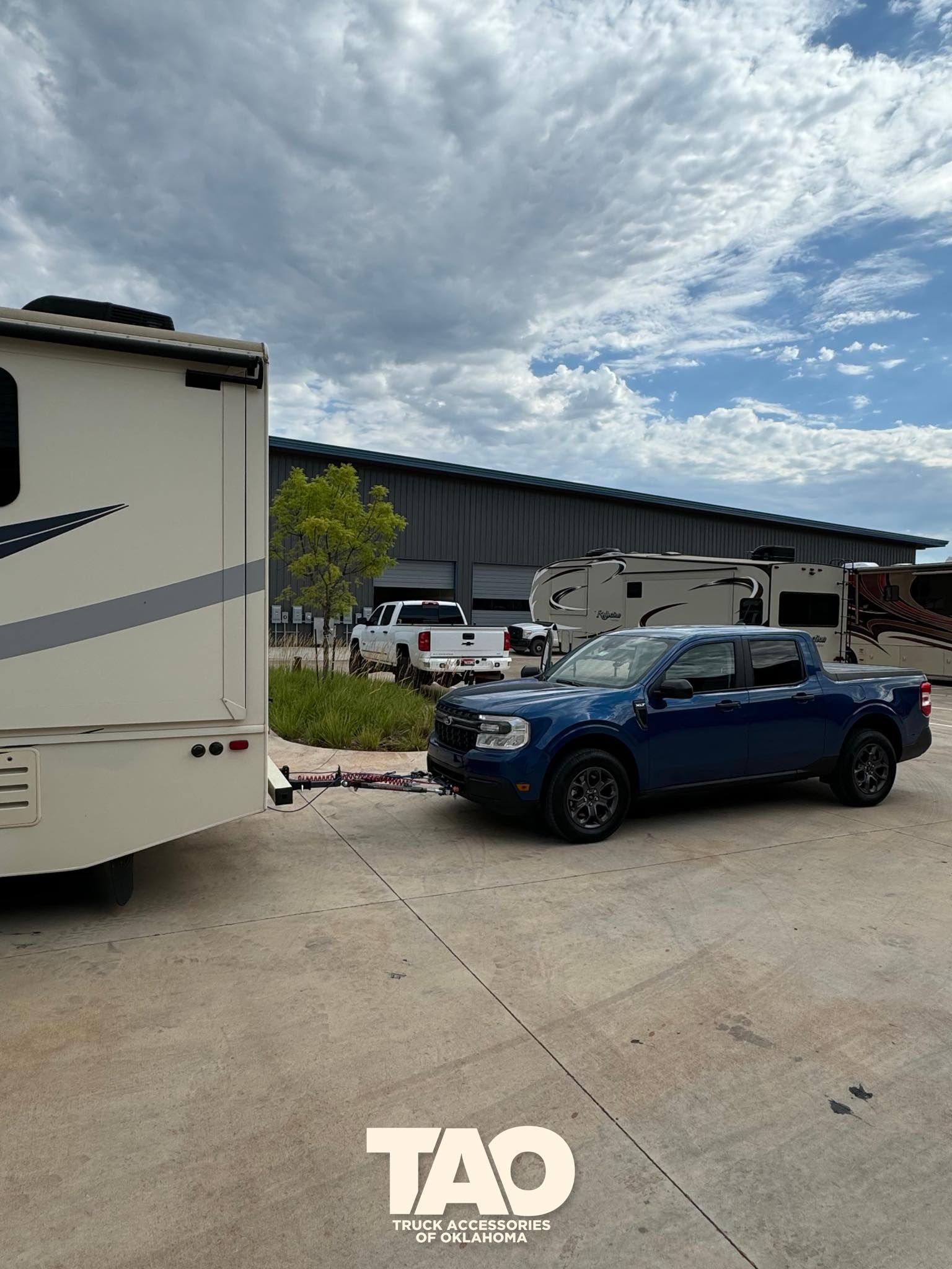 Blue Ford pickup truck towing an RV on a paved lot with cloudy sky.