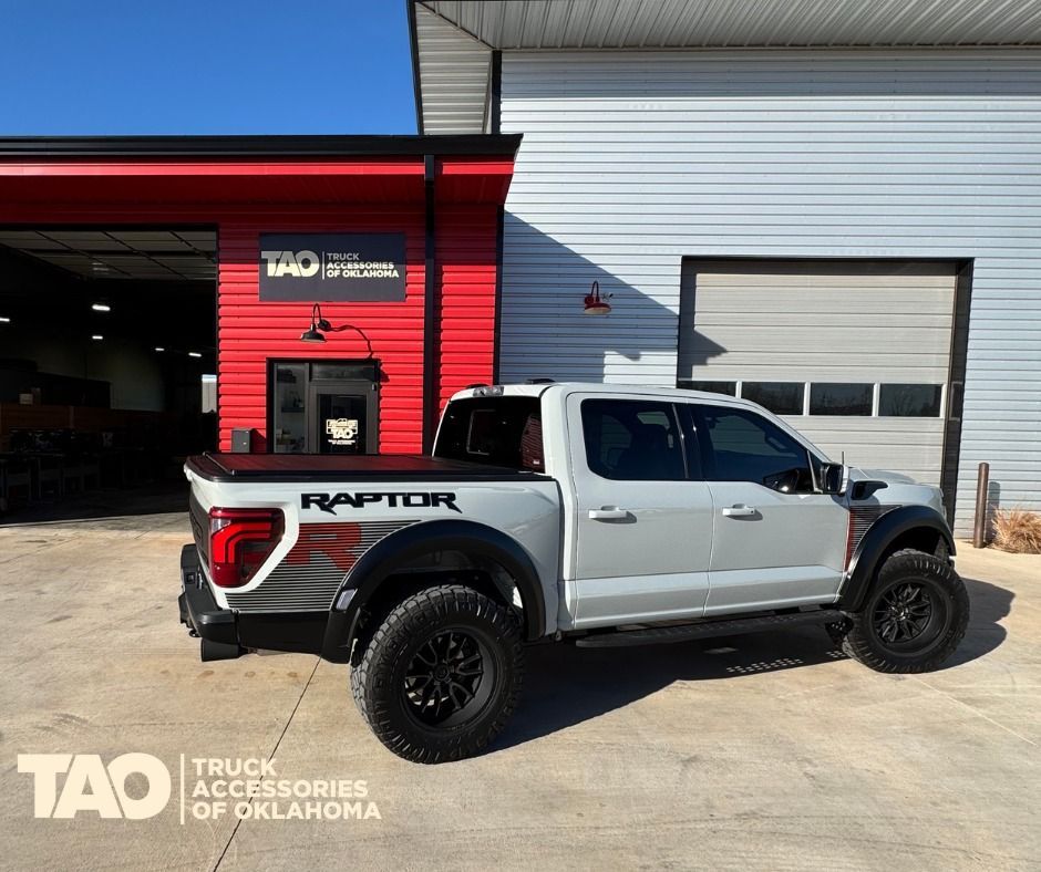 Silver Ford Raptor truck parked outside a red and gray building with black tires.