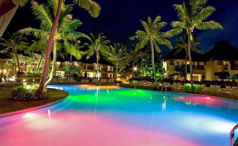 A large swimming pool surrounded by palm trees is lit up at night.
