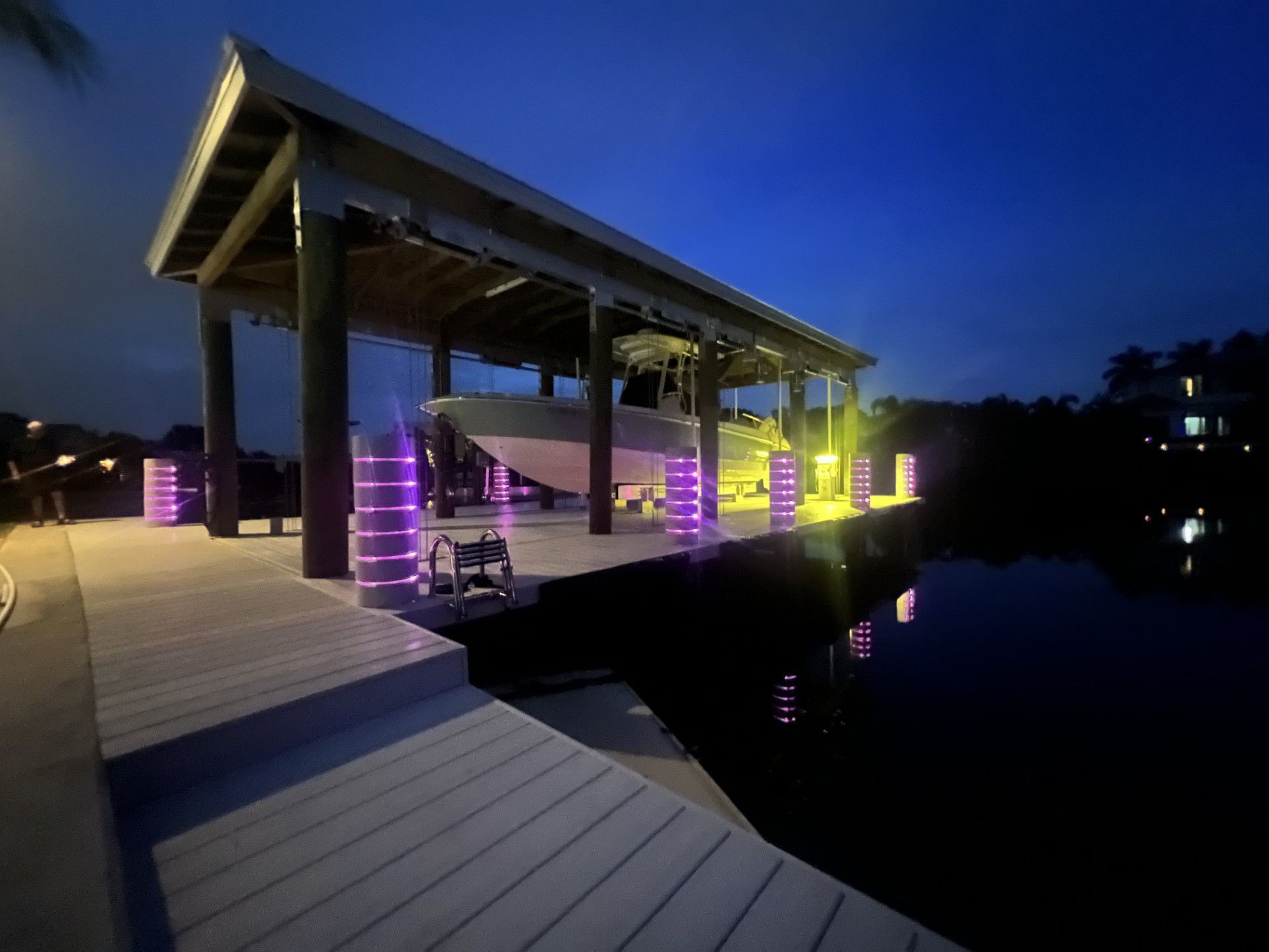 Dock at night with a boat under a covered structure, illuminated by purple and yellow lights. 