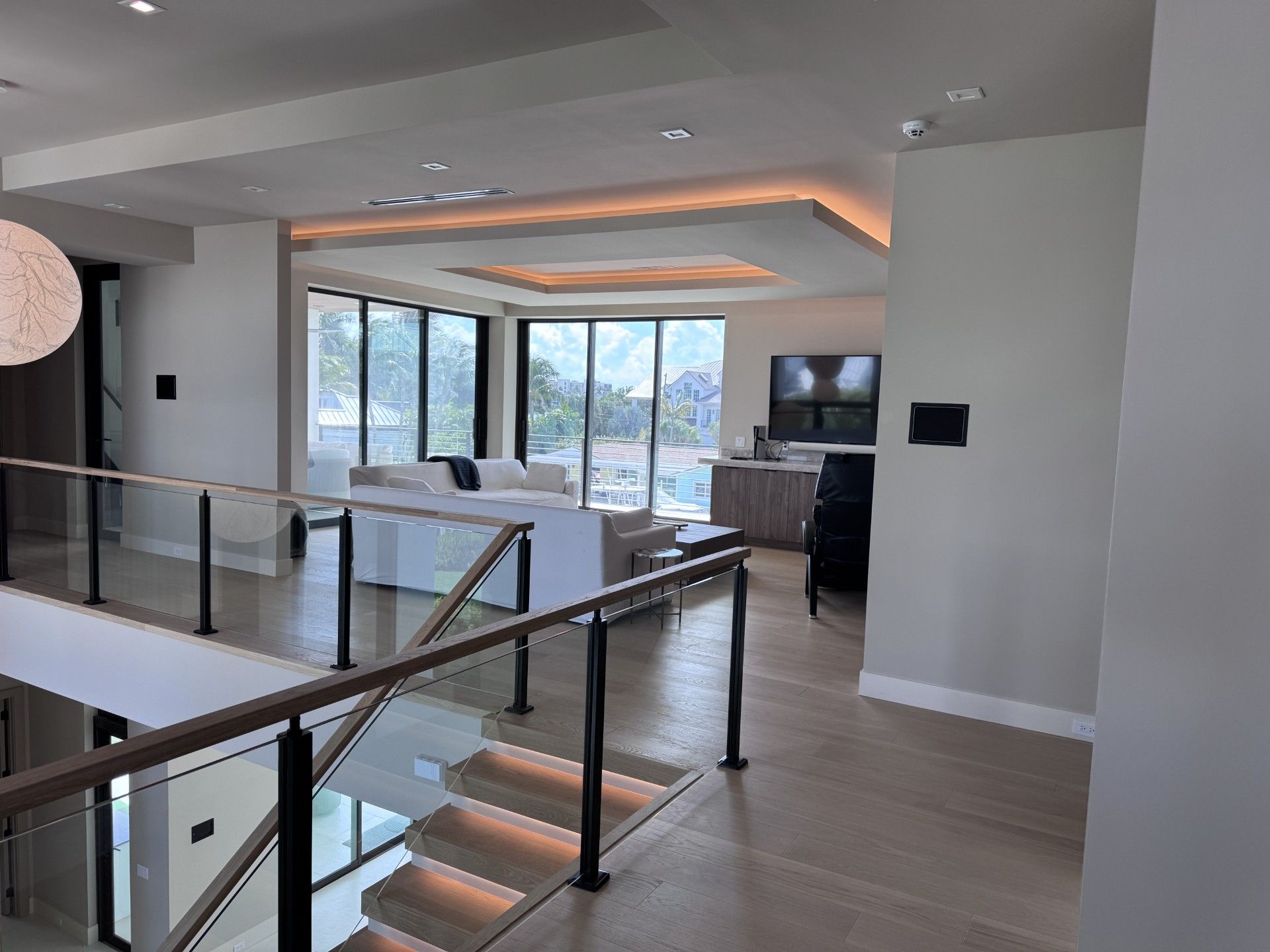 Modern home interior: foyer with staircase, glass railing, and living area with large windows, sofa, and a television. 