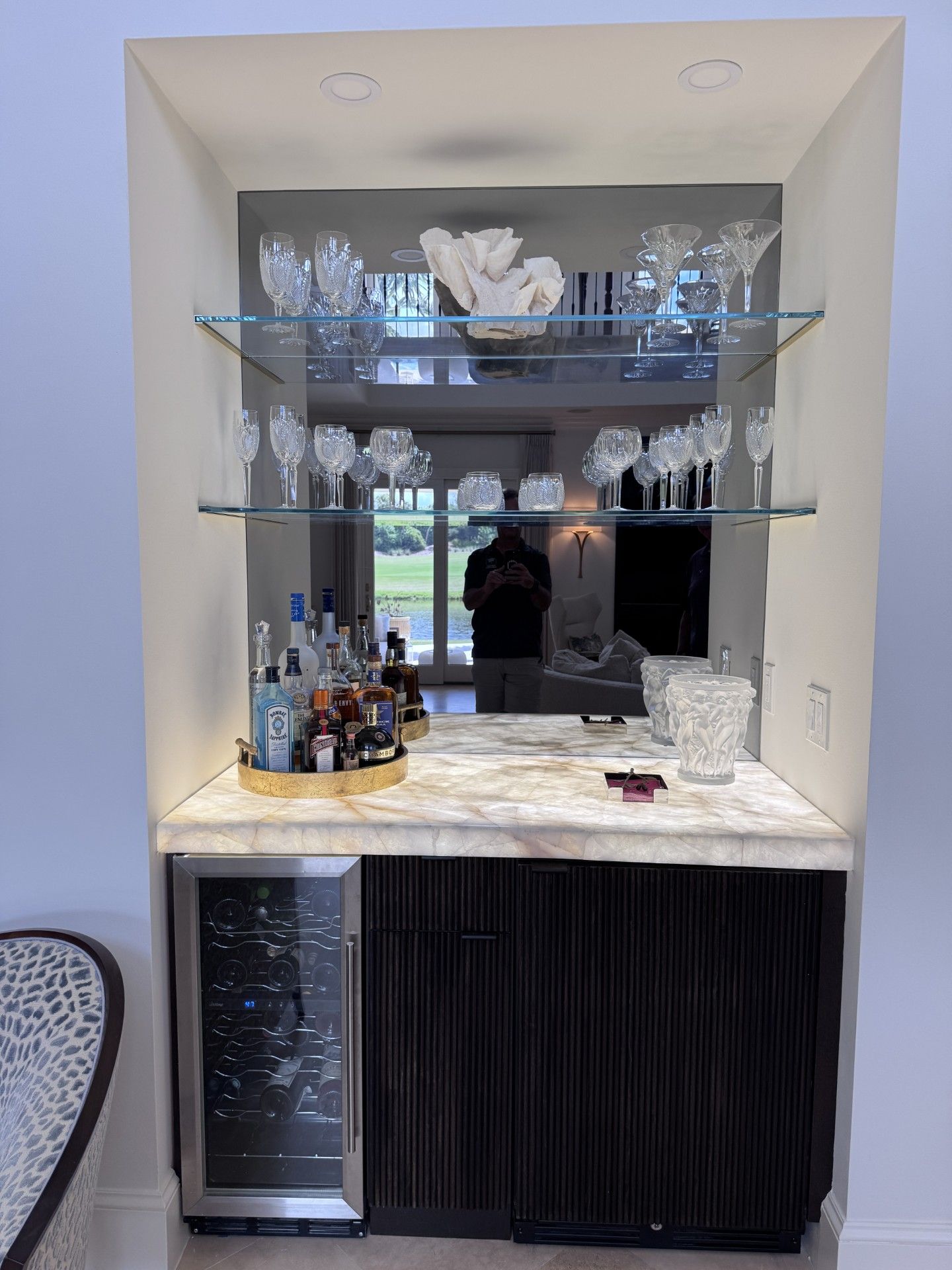 Built-in bar with glass shelves, a mirrored back, and a mini-fridge. Displays glassware, liquor bottles, and a countertop. 