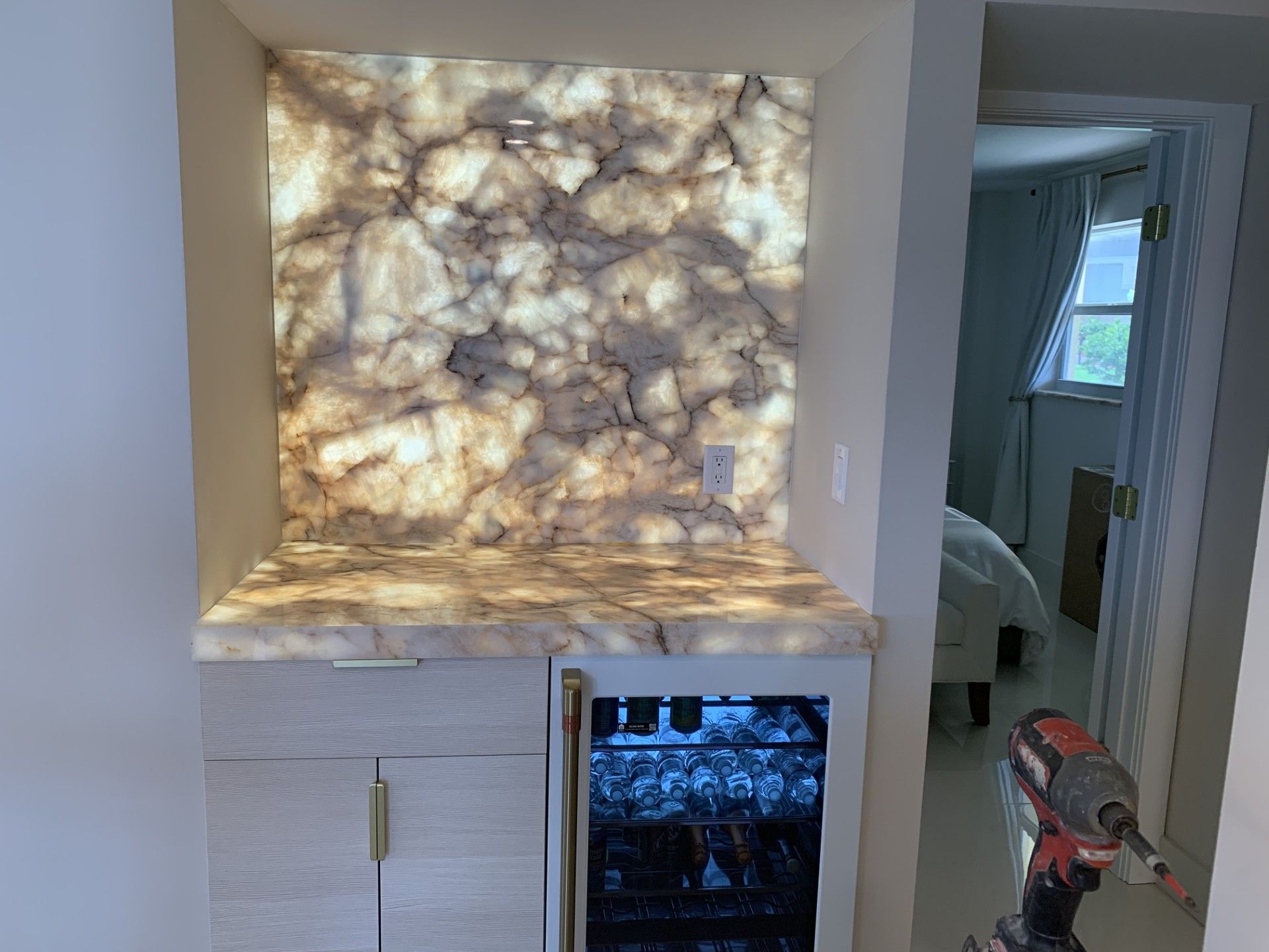 Built-in bar with illuminated onyx backsplash and countertop, wine fridge, and cabinetry. Doorway to a bedroom. 