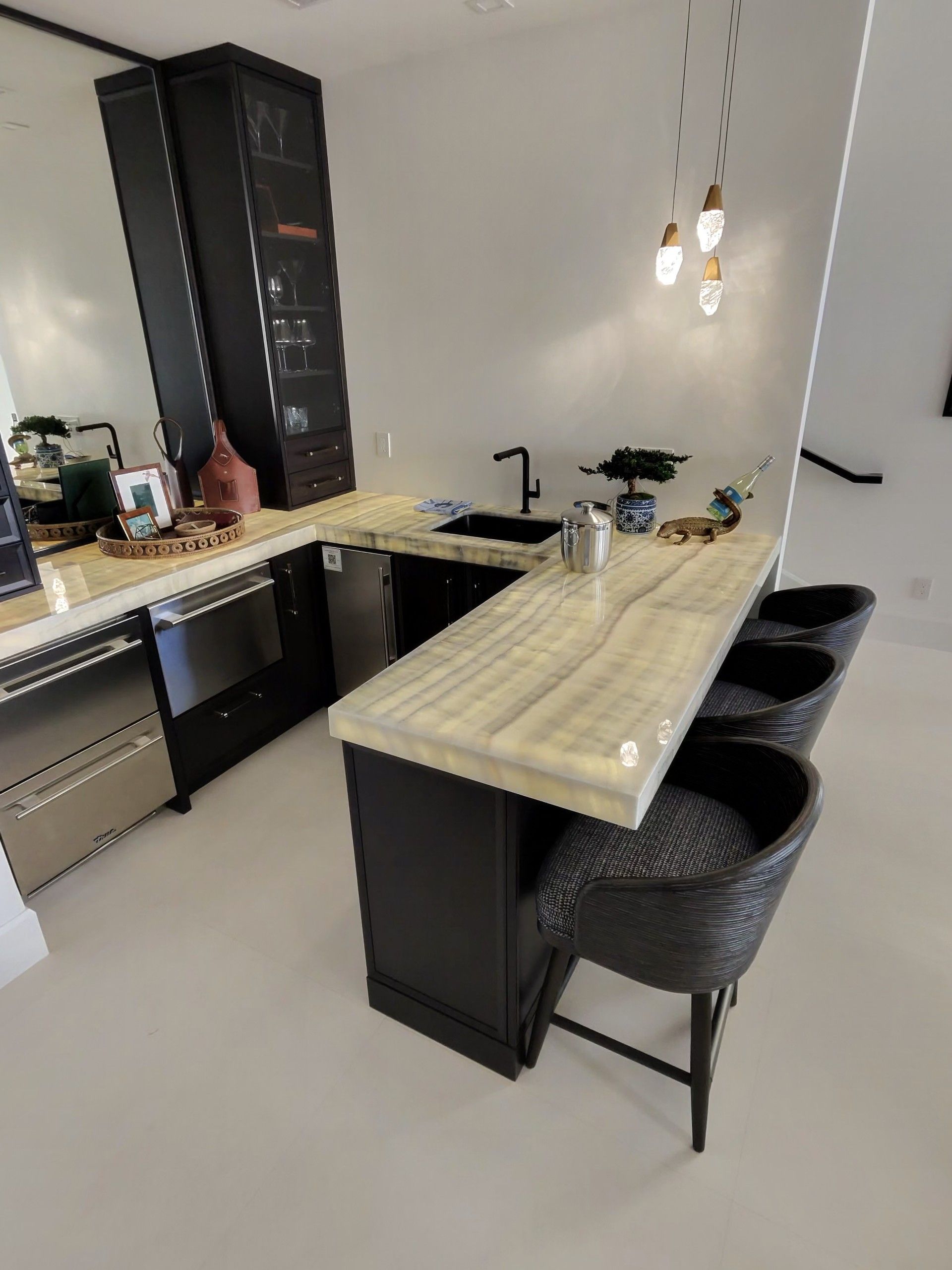 Modern home bar with black cabinetry, backlit marble countertop, black stools, and pendant lights. 