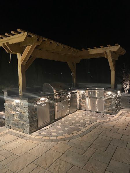 A large outdoor kitchen with stainless steel appliances under a pergola at night.