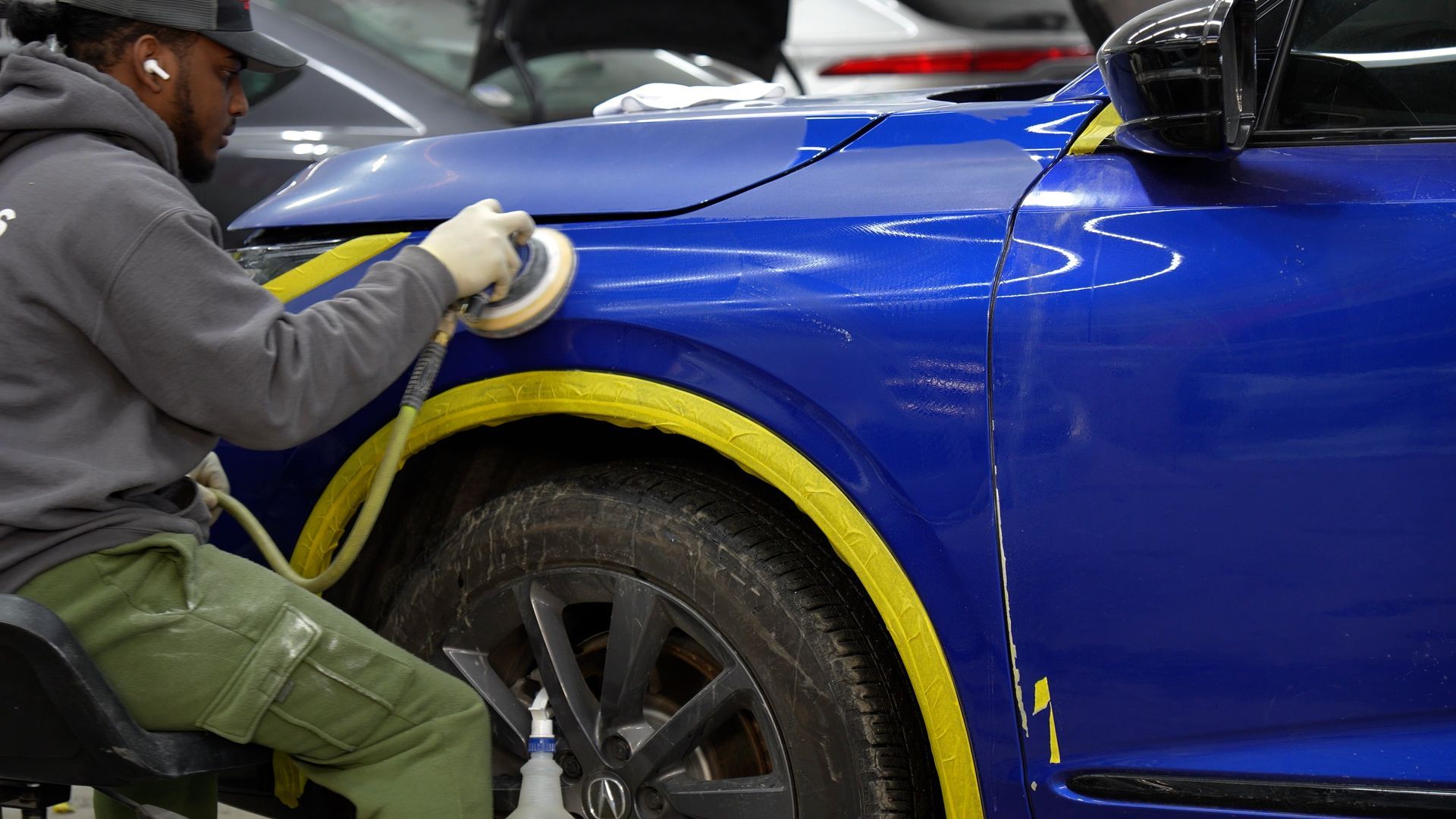 Worker polishing a blue car door with a buffer, masking tape around the wheel well in an auto shop.