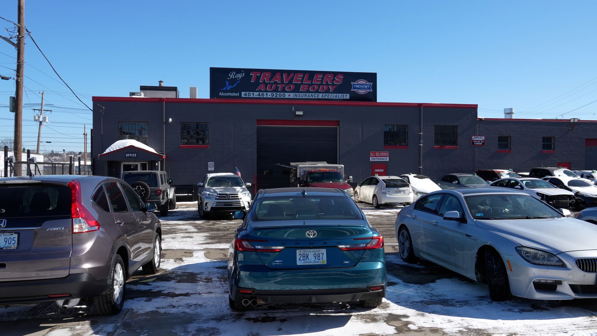 Parking lot outside a gray auto repair shop with several cars and patches of snow on the ground.