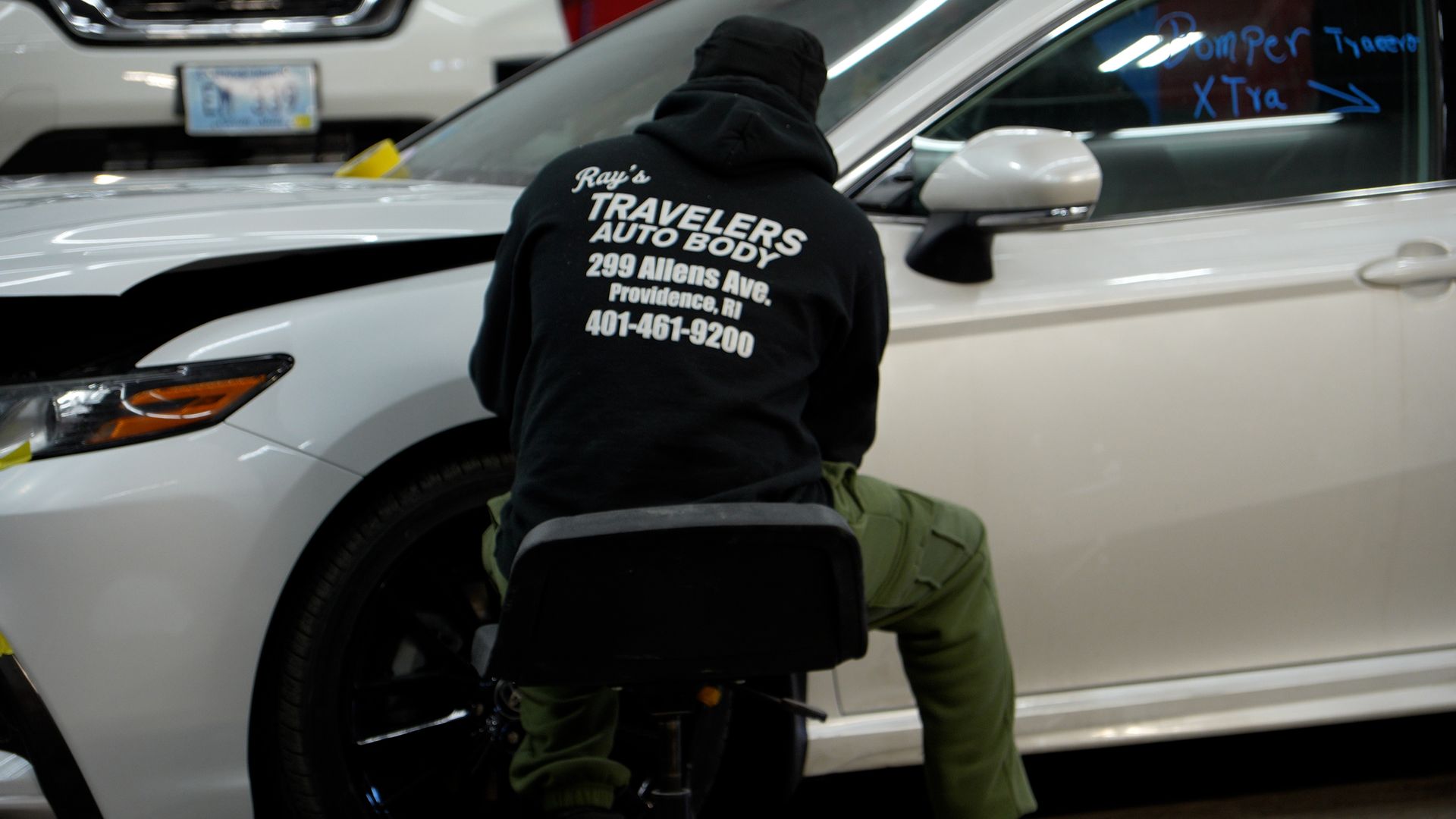 Person in a black jacket crouches by a white car, likely working on the front wheel.