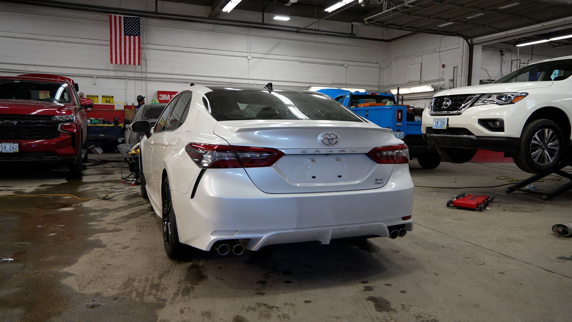 White sedan in an auto repair shop with lifted vehicles and tools on the floor.