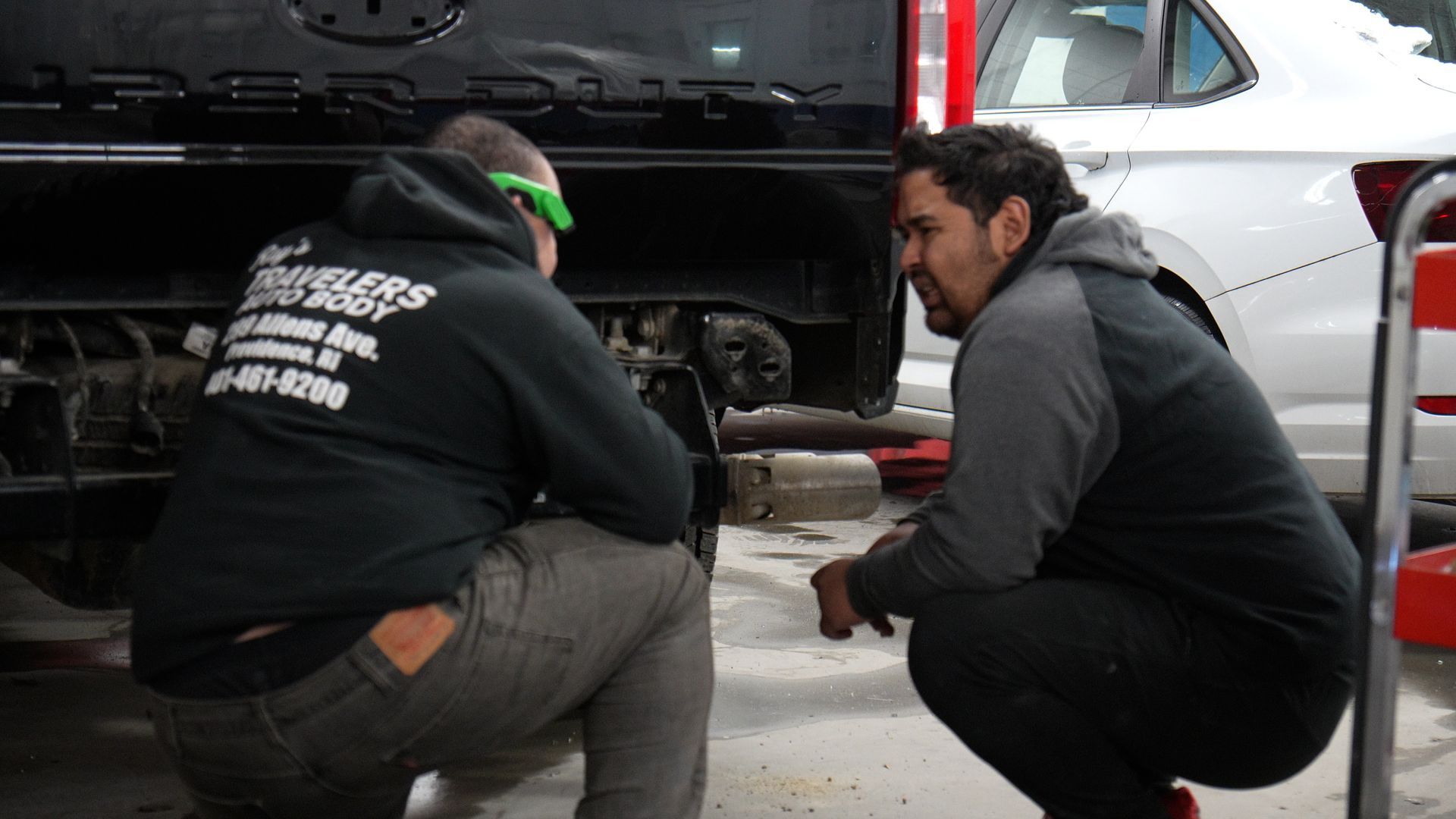 Two mechanics crouch beside a black vehicle, inspecting the underside near the rear wheel.
