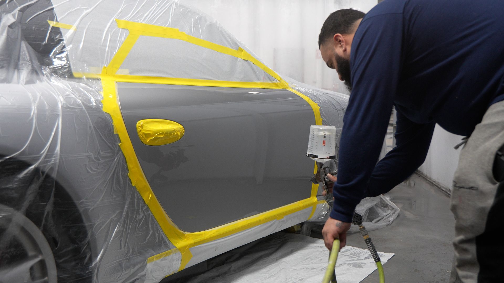 A man in blue work clothes applies masking tape to a gray car door in a repair shop.