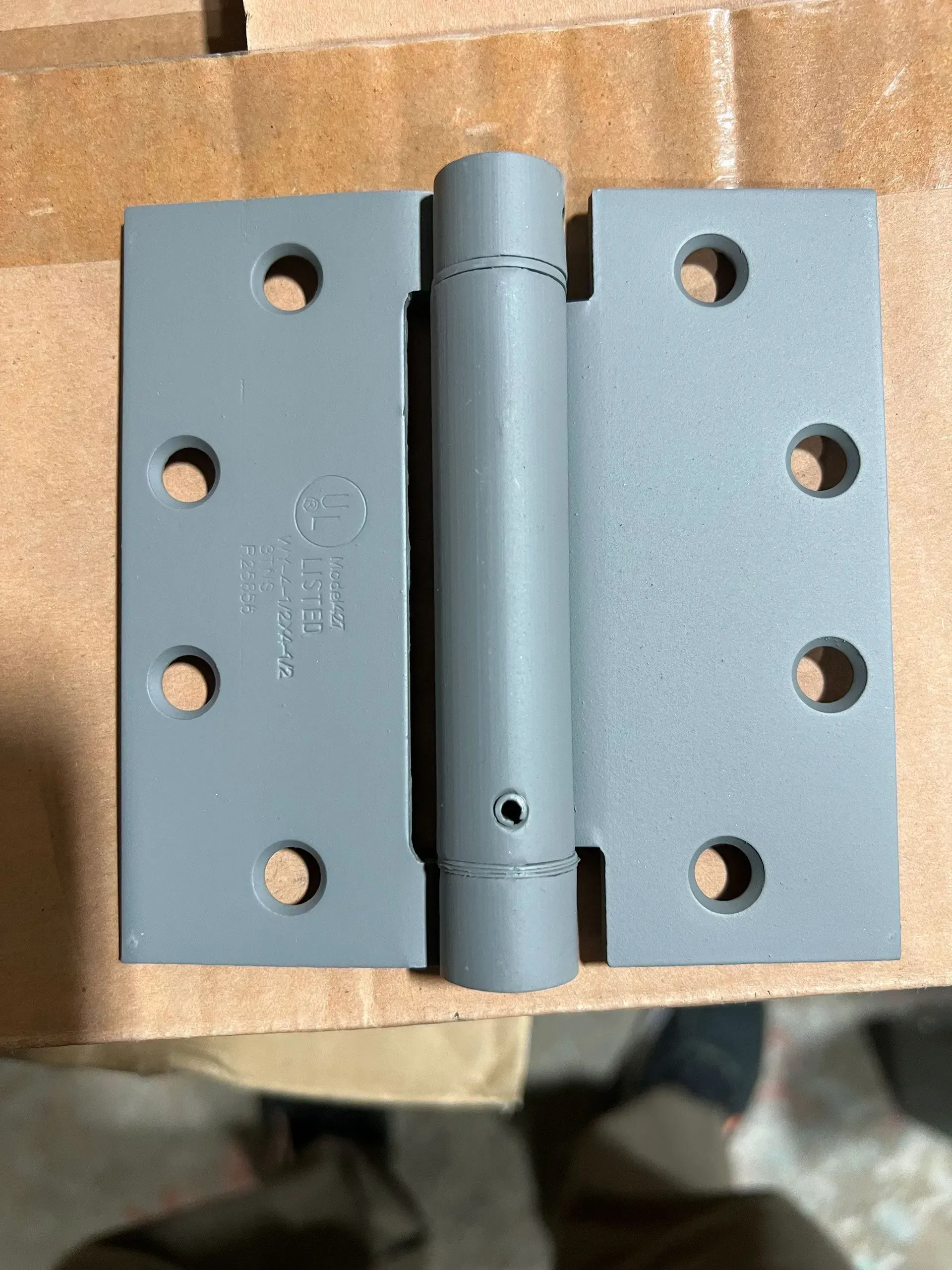 Gray spring hinge with six screw holes on each leaf, sitting on cardboard.