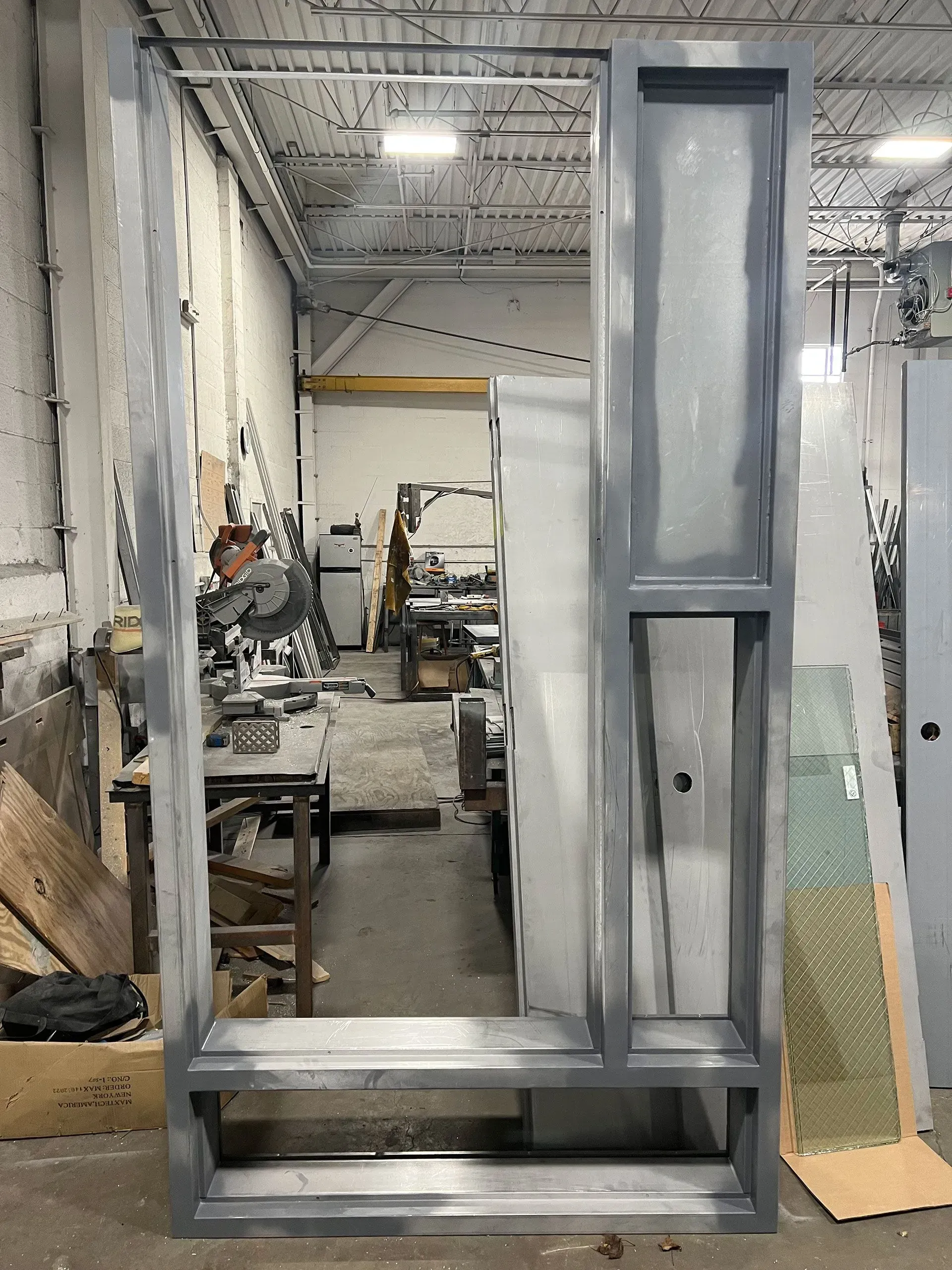 Metal framed structure, likely for a door or window, in a workshop setting. Gray painted frame, with an open interior view.
