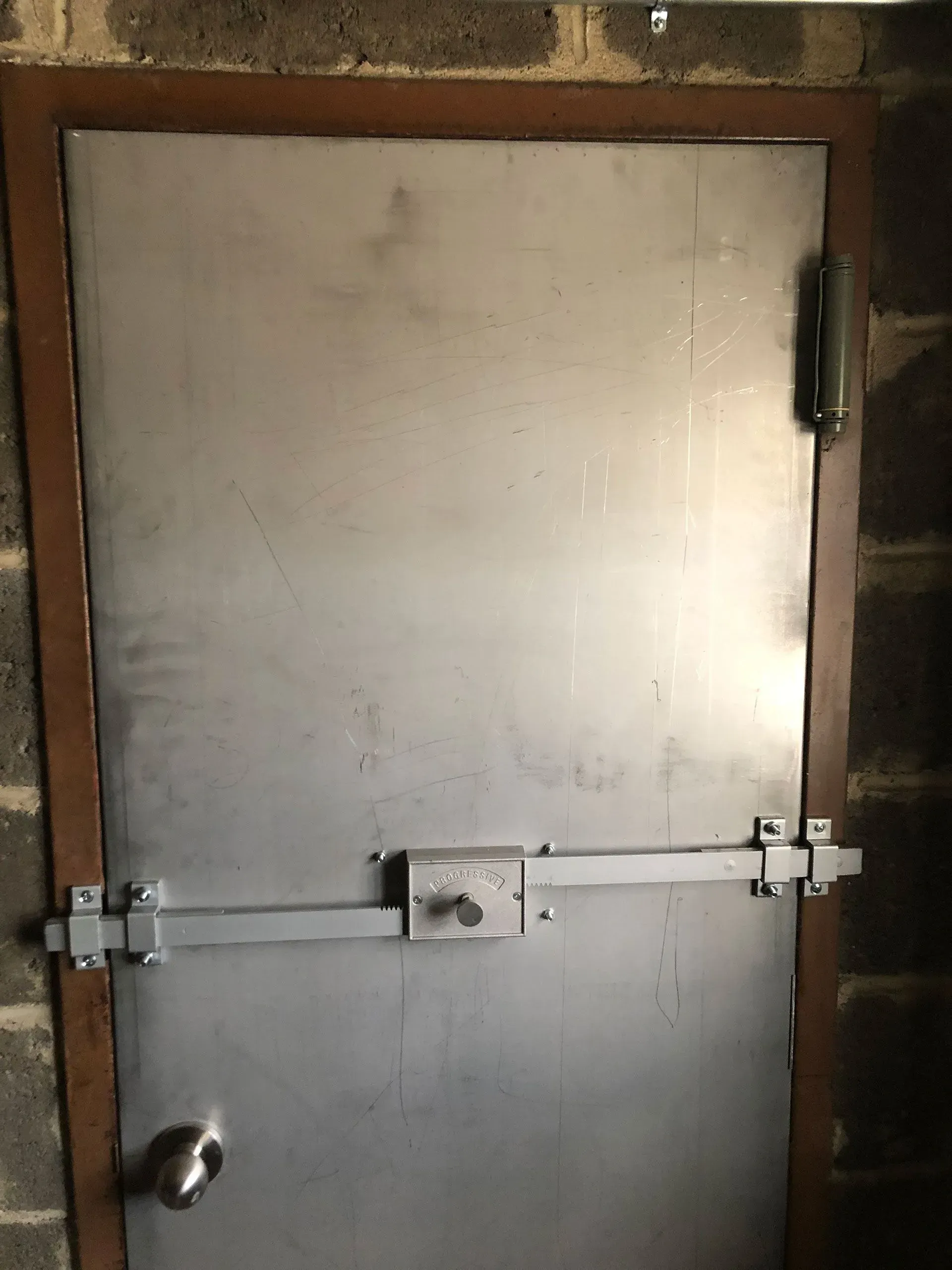 Metal door with a simple locking mechanism, set in a brown frame against a brick wall.