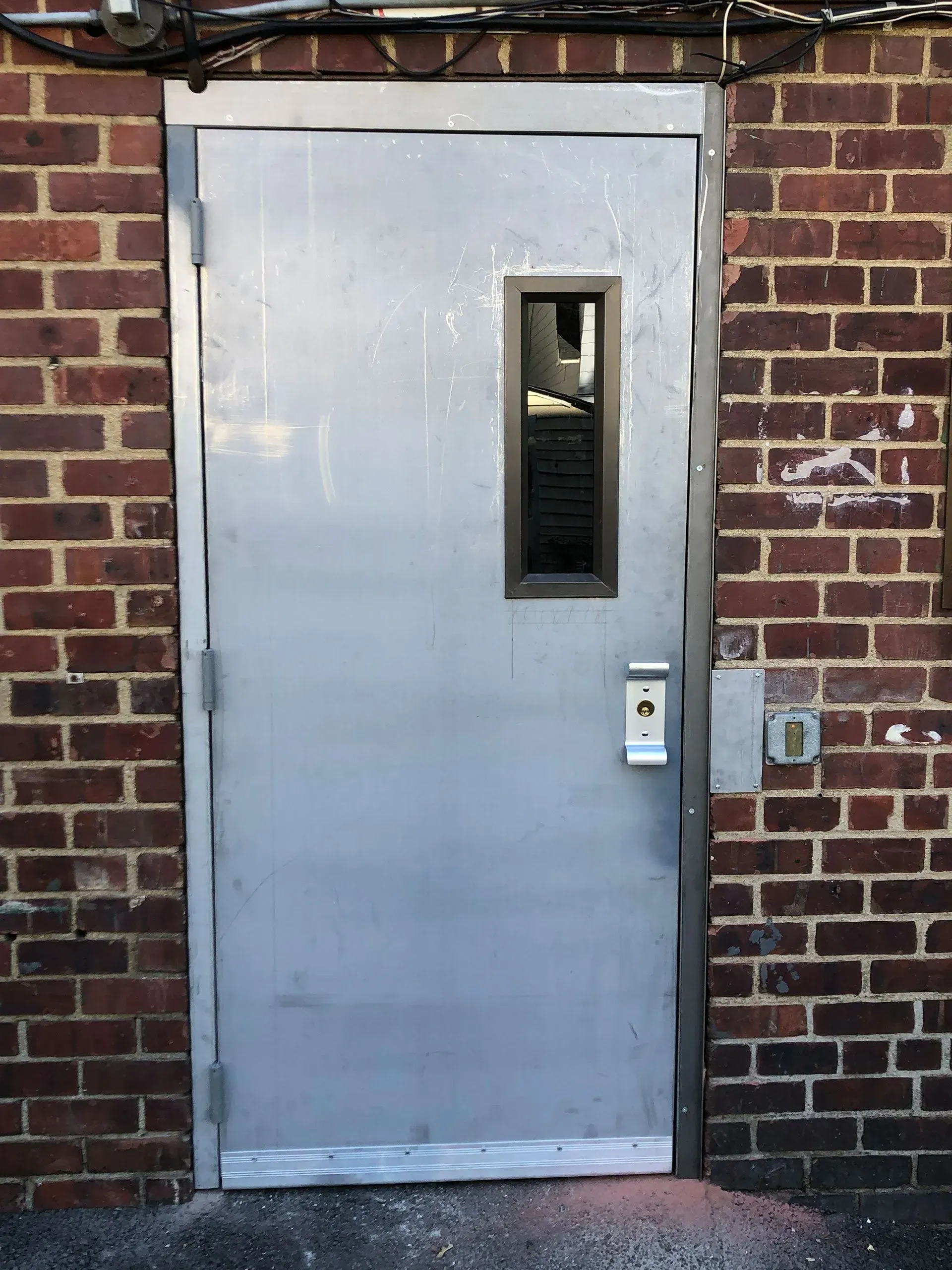 Silver metal door with a rectangular window set in a brick wall.