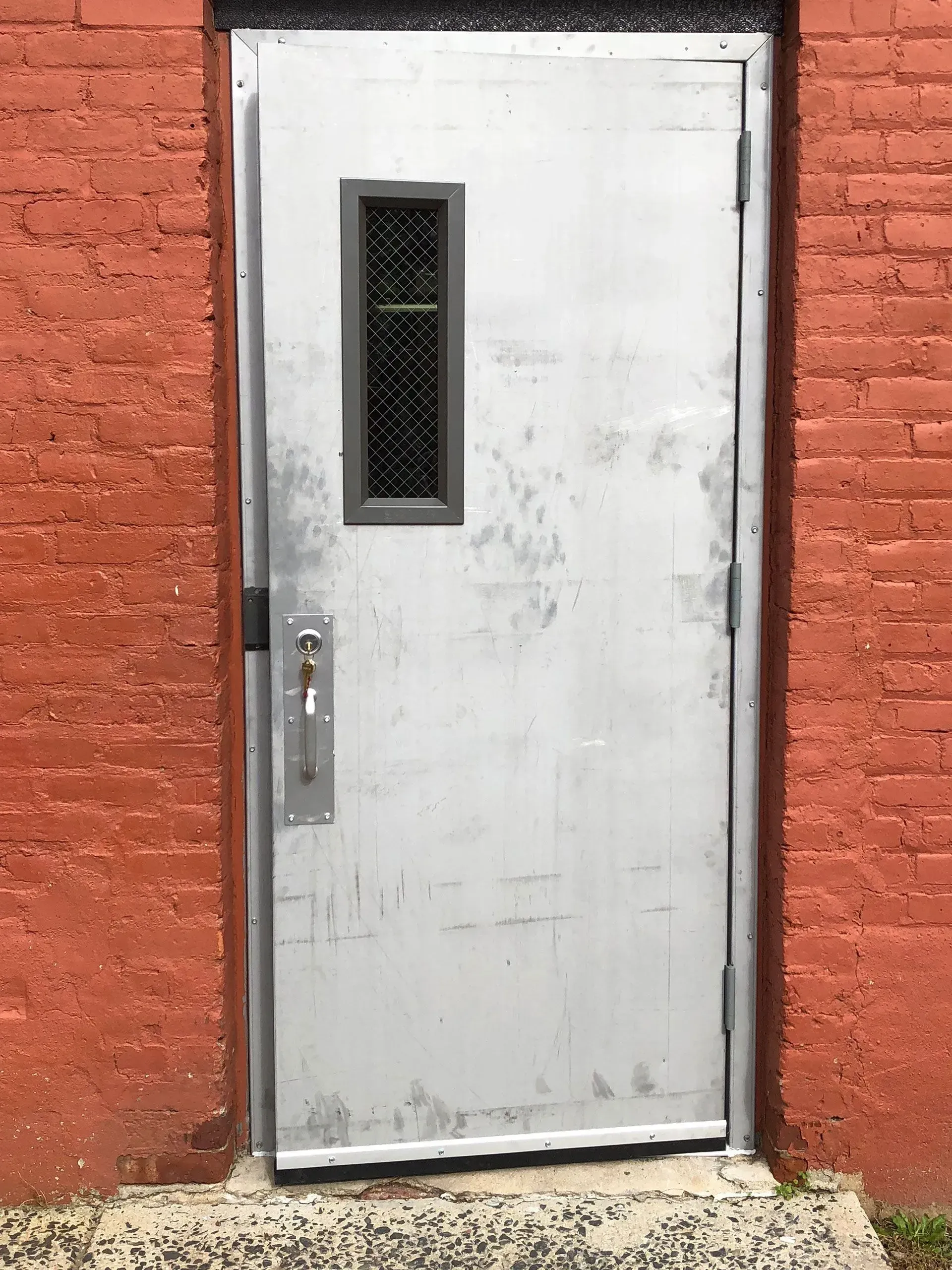 Gray metal door with small window, set in red brick wall.
