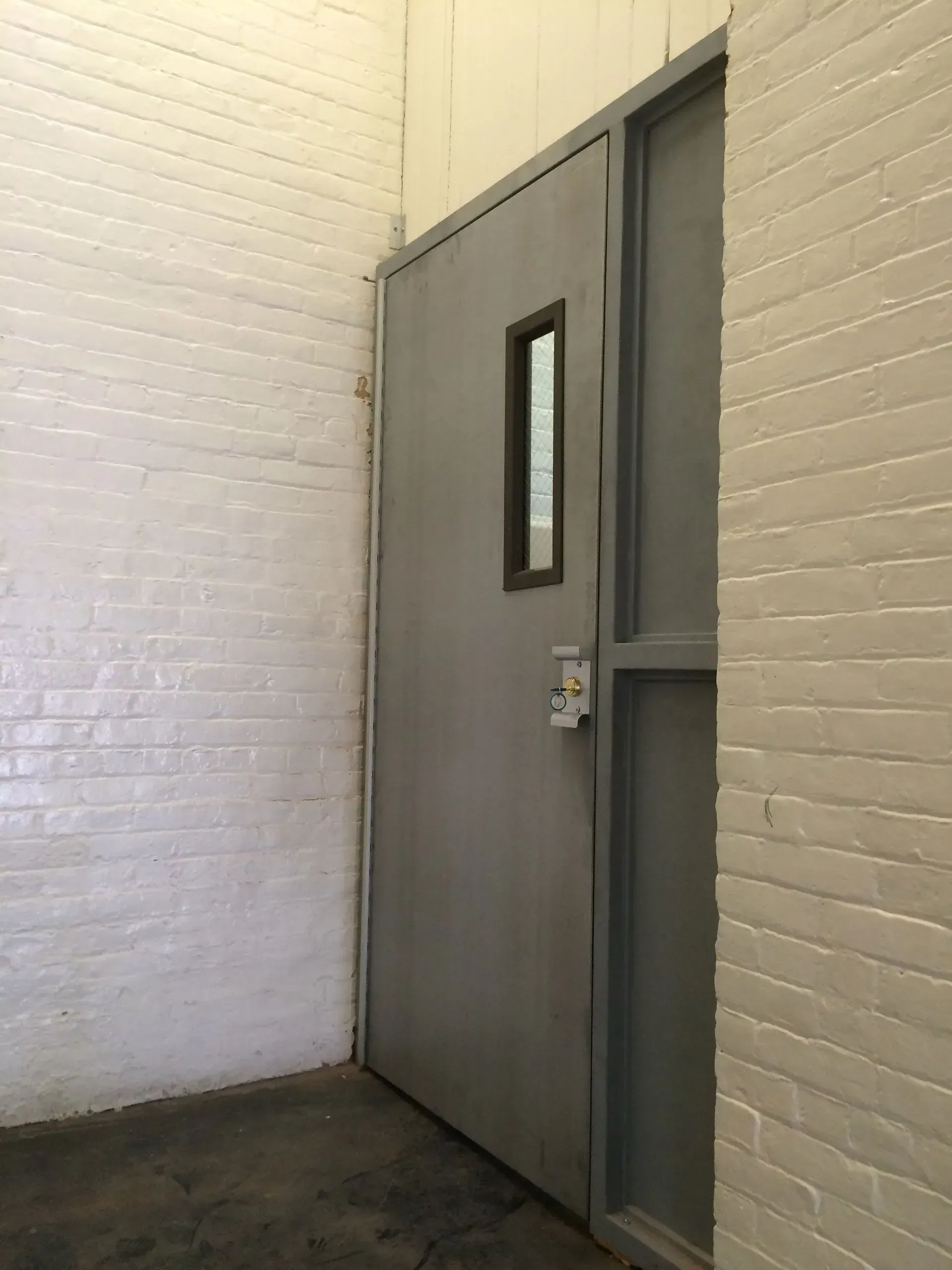 Gray metal door with small window, set in white brick wall, in an indoor setting.