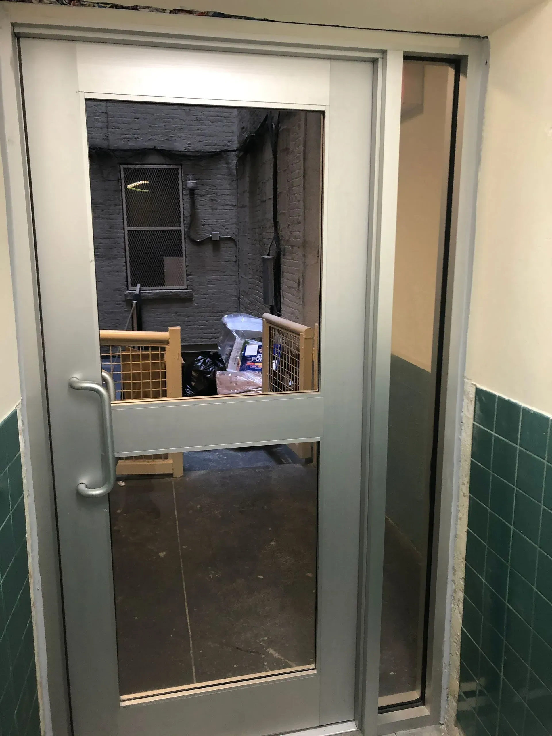 Glass door with silver frame, looking into an alley. Brick walls are visible, as are a couple of wooden chairs inside.