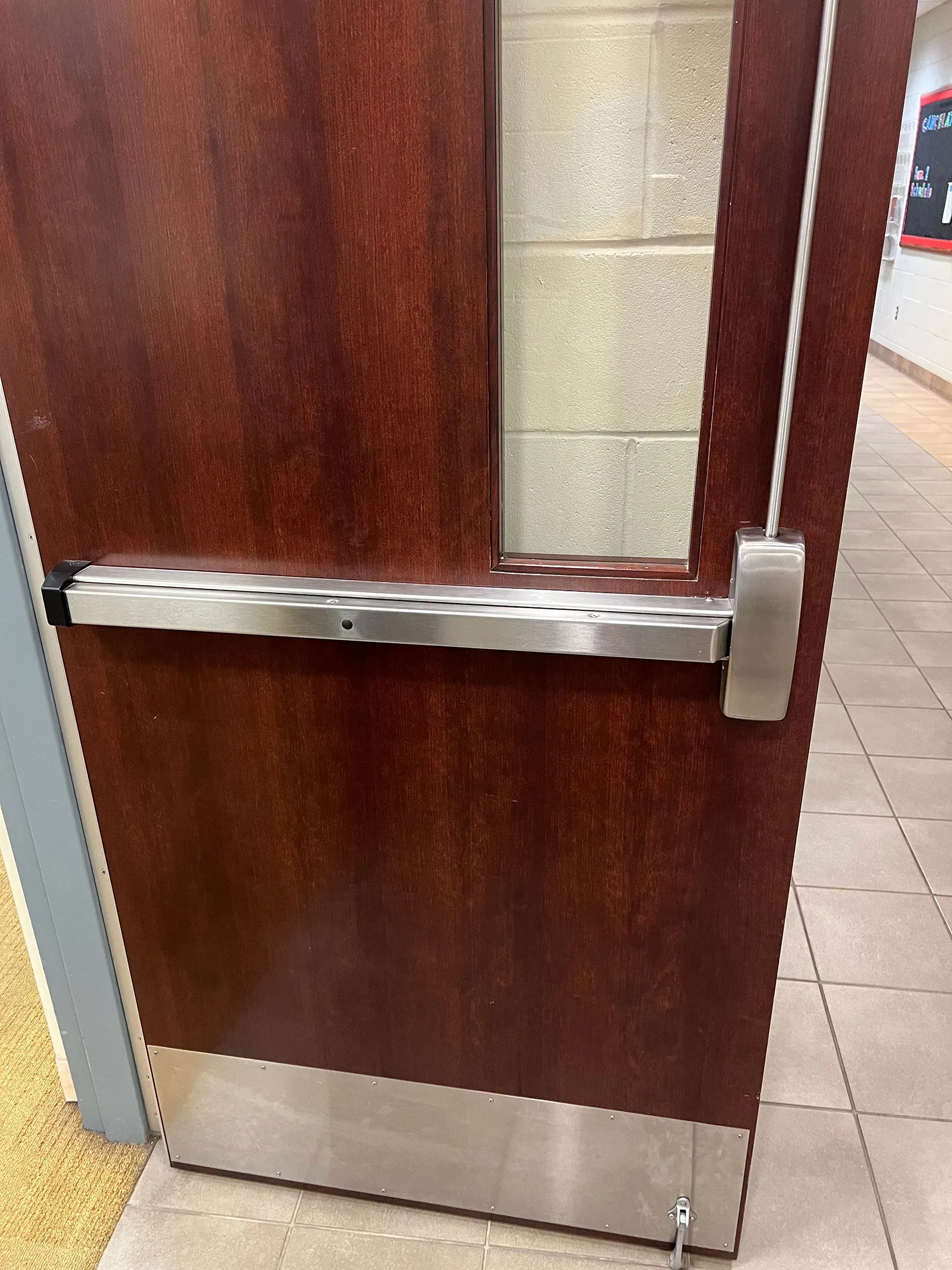 Wooden door with a metal push bar and kick plate; a hallway is visible.