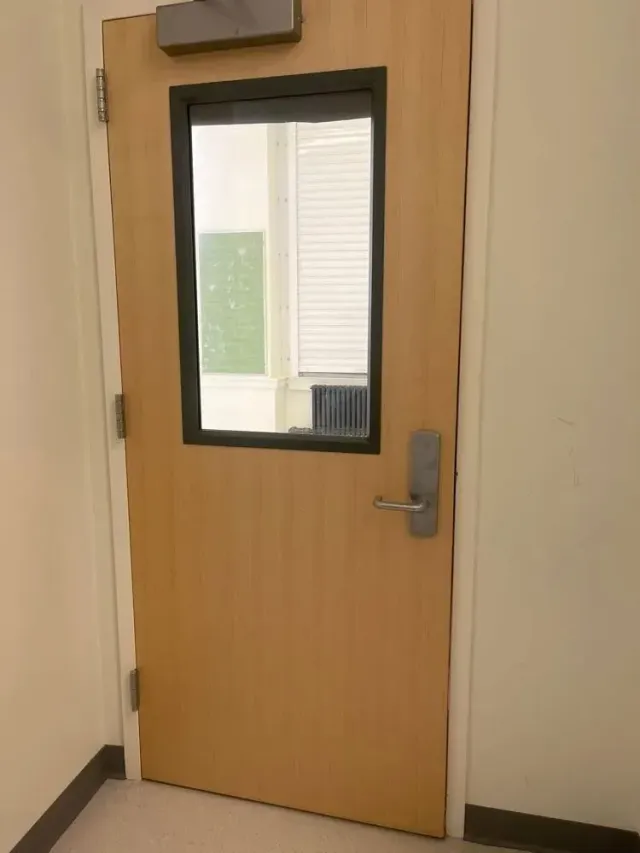 Tan door with rectangular window, metal handle, and closer. Light beige walls and baseboard visible.
