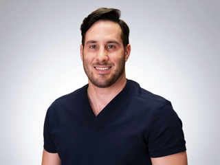 A person with a beard, wearing a dark blue v-neck scrub top, smiling against a plain, light gray background.