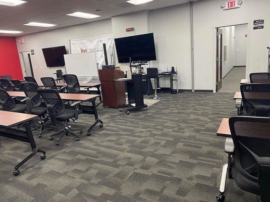 A carpeted basement with tables and chairs and a television on the wall.