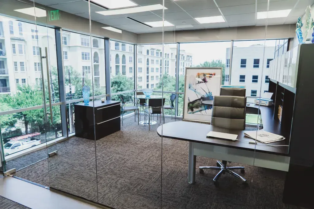 An office with a desk and chairs in front of a glass wall.