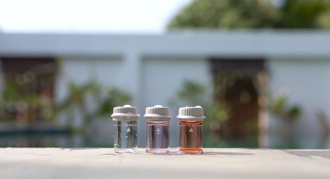 Three small, clear bottles with colored liquids, outdoors against a blurry white wall and greenery.
