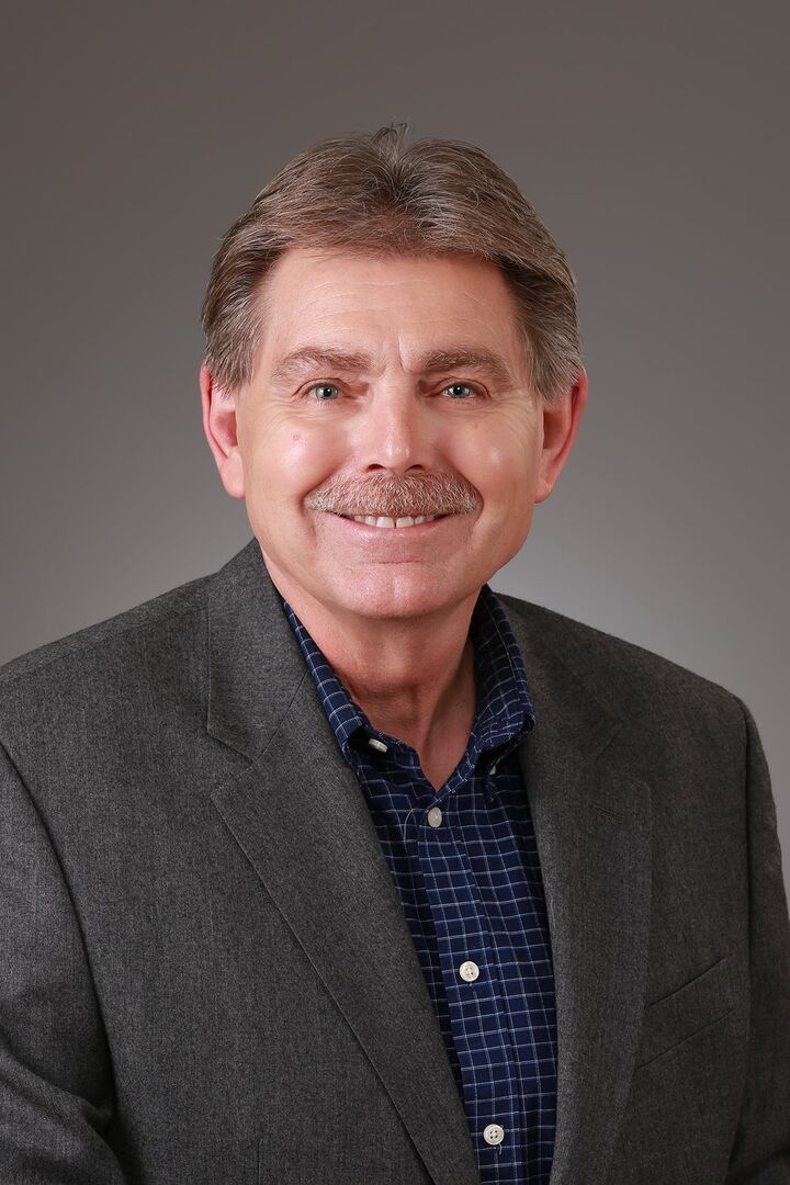 Smiling man in a gray blazer and dark shirt against a plain gray background
