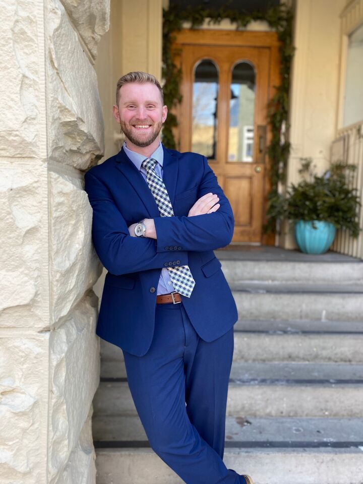 Man in a blue suit leaning against a stone wall on steps, smiling in front of a wooden door