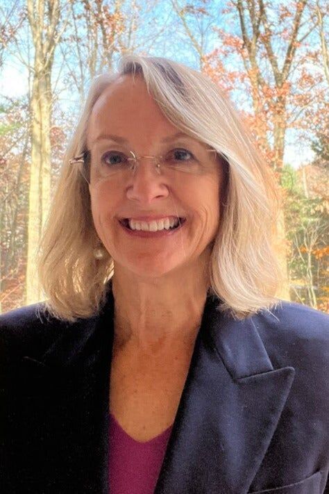 Smiling person with blonde hair and glasses, wearing a dark blazer outdoors with autumn trees behind.