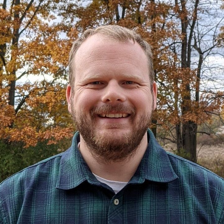 Smiling man in a blue plaid shirt outdoors with autumn trees in the background