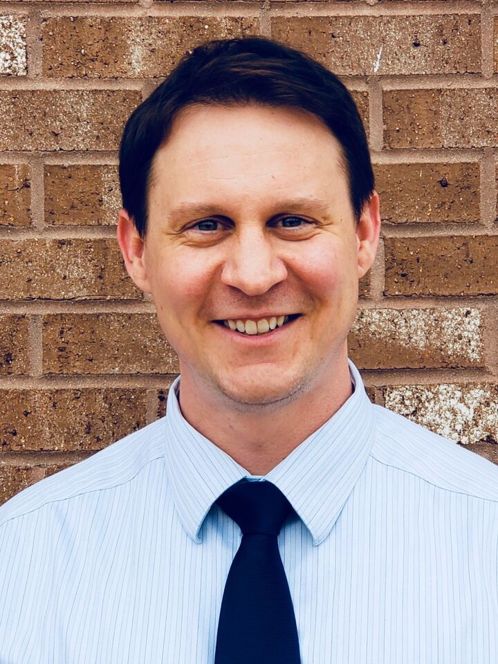 Smiling man in a light blue shirt and navy tie standing in front of a brick wall