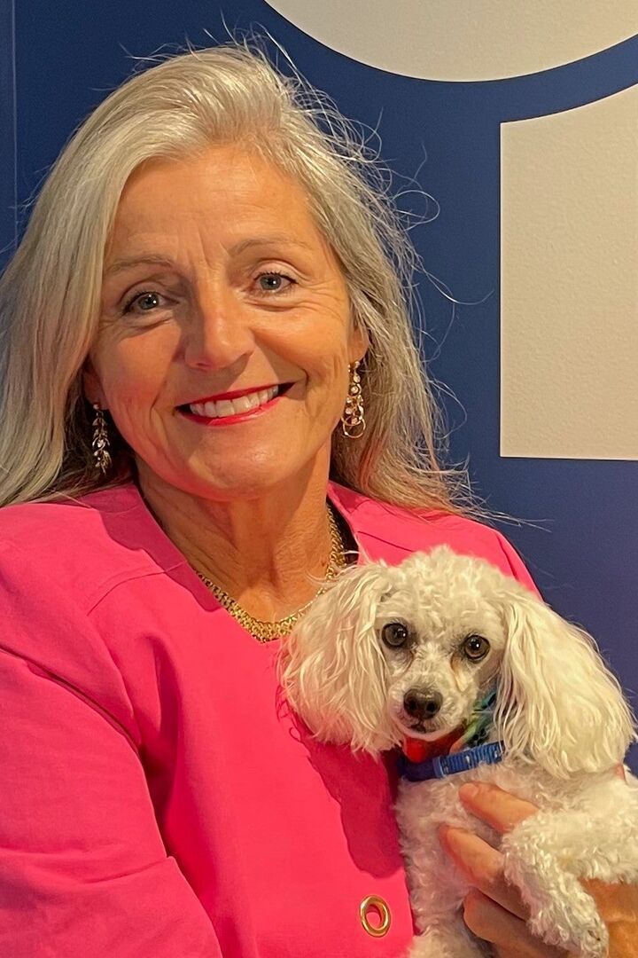 Smiling woman in a pink jacket holding a small white dog against a blue backdrop